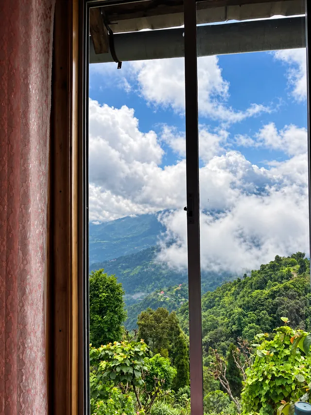 Green hills and billowing clouds seen through a wooden-framed window with a pink curtain